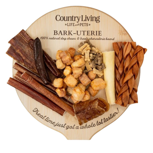 Country Living Bark-Uterie Board dog treat assortment with bully sticks, gullet flats, cow tail, chicken sausages, beef trachea, braided collagen sticks, yak chew puffies, and soft chew biscuits arranged on a rustic wood-look cardboard board.